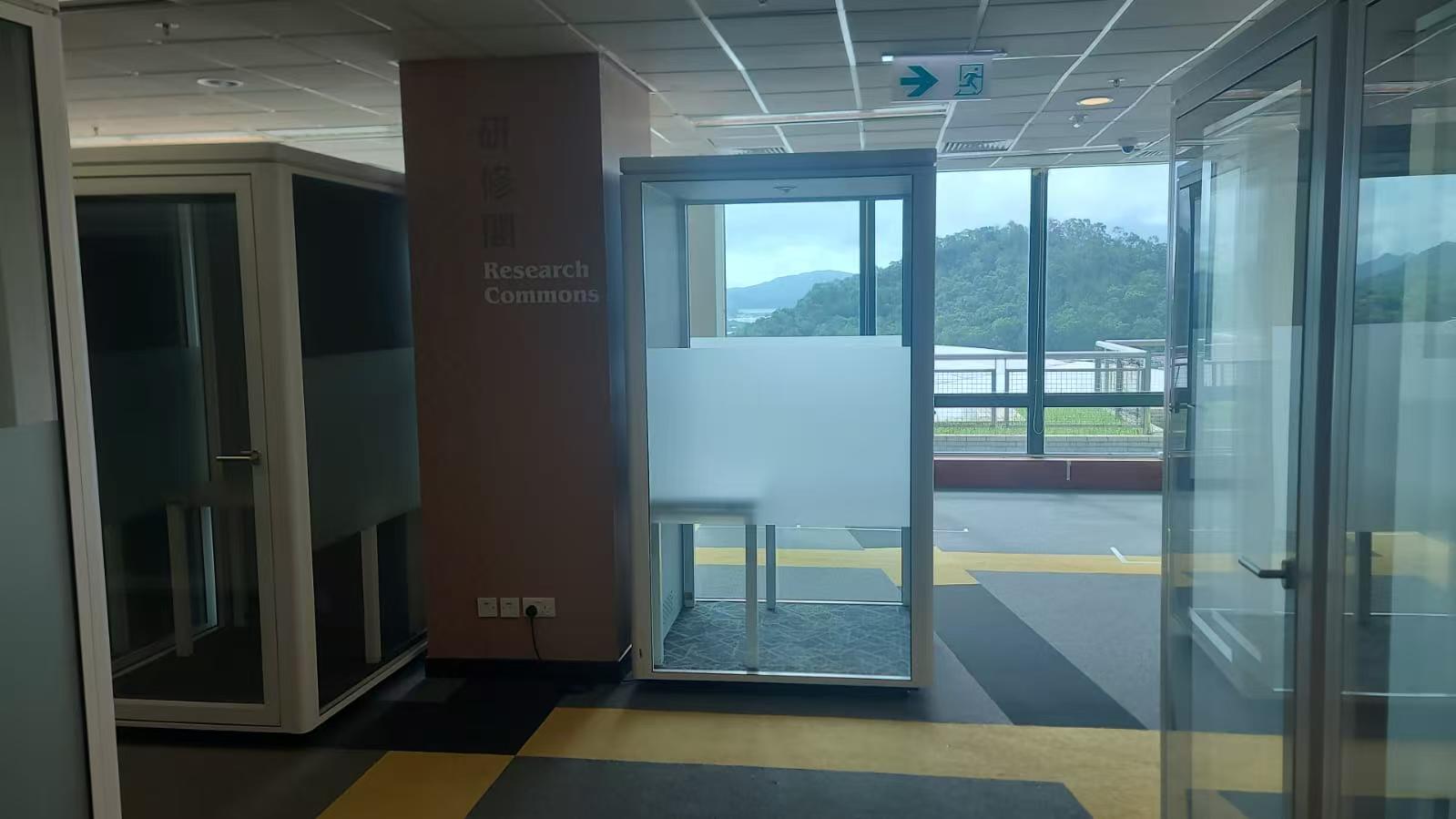 An office interior with glass-walled cubicles and a large window overlooking a green outdoor landscape. A brown pillar on the left has the words 'Research Commons' printed on it.