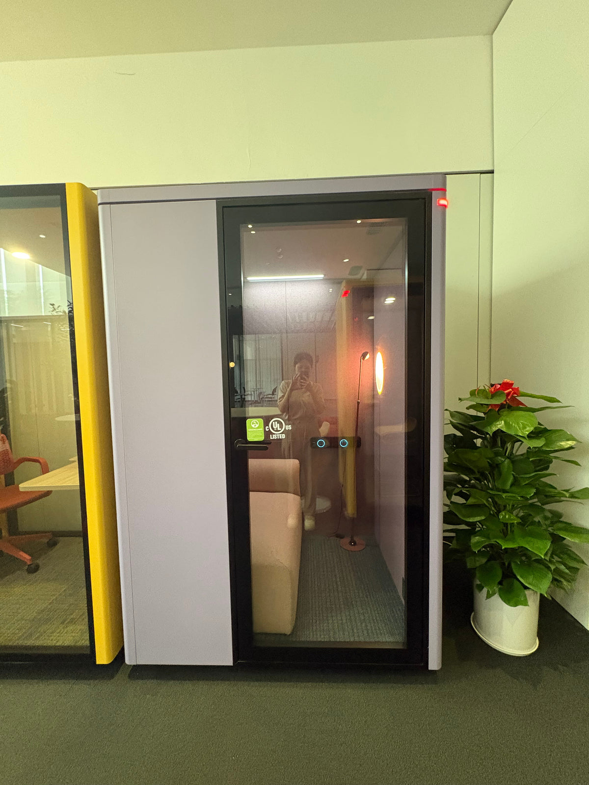 A modern glass office phone booth with a white side panel and black frame, shown in an office setting next to a yellow partition wall and a potted plant.