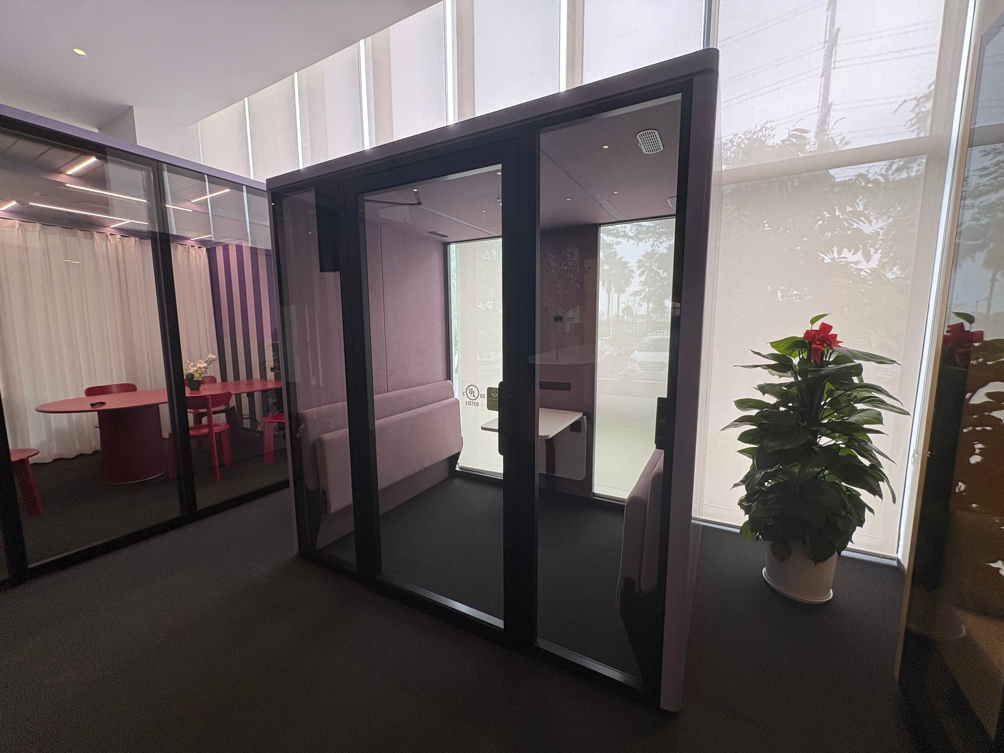 A modern glass partition wall with a black metal frame, shown installed in an office interior with a plant nearby and other office spaces visible through mirrored glass.