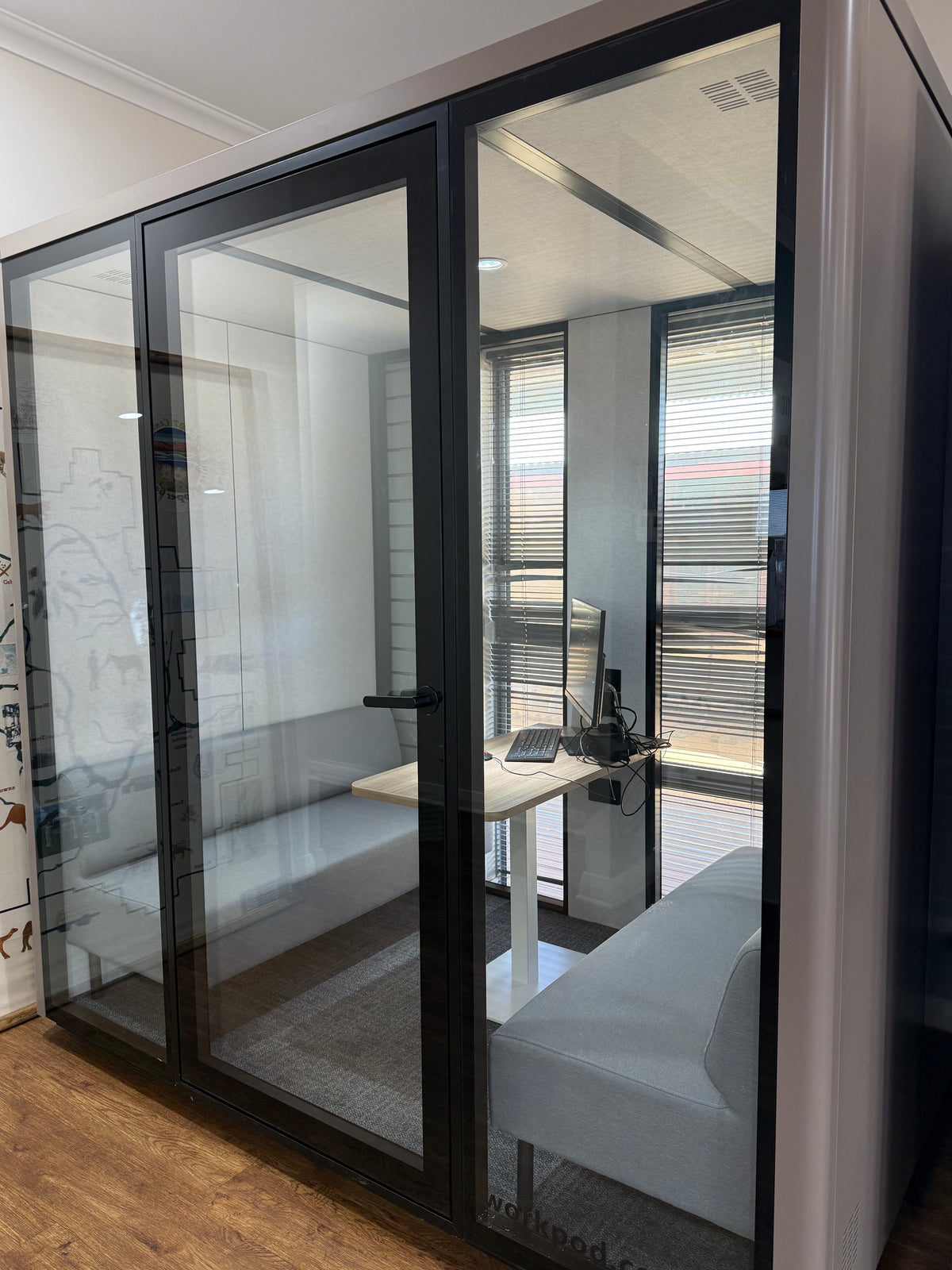A modern glass office partition pod with black metal frames, shown installed in an office space with a desk, computer, and seating visible inside.