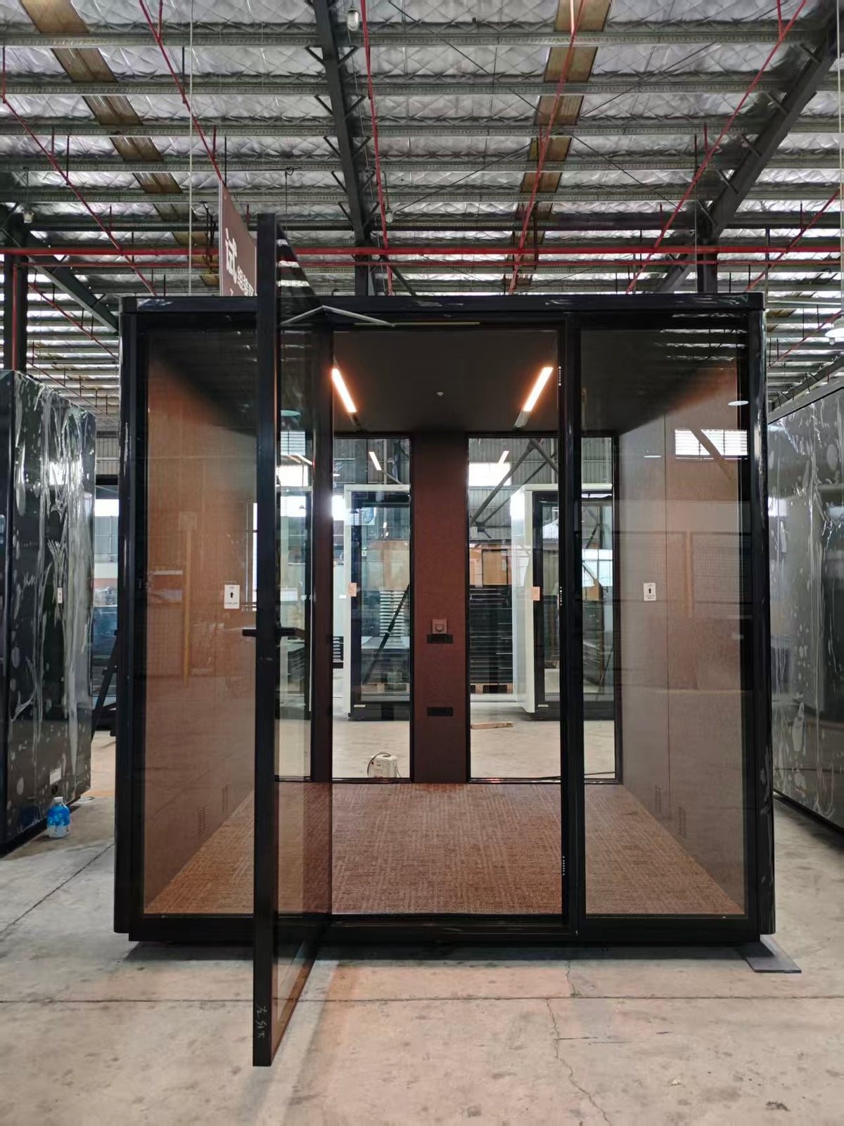 A modern outdoor cabana or small cabin with a black metal frame, glass walls, and a brown woven mat floor, shown in a warehouse setting.