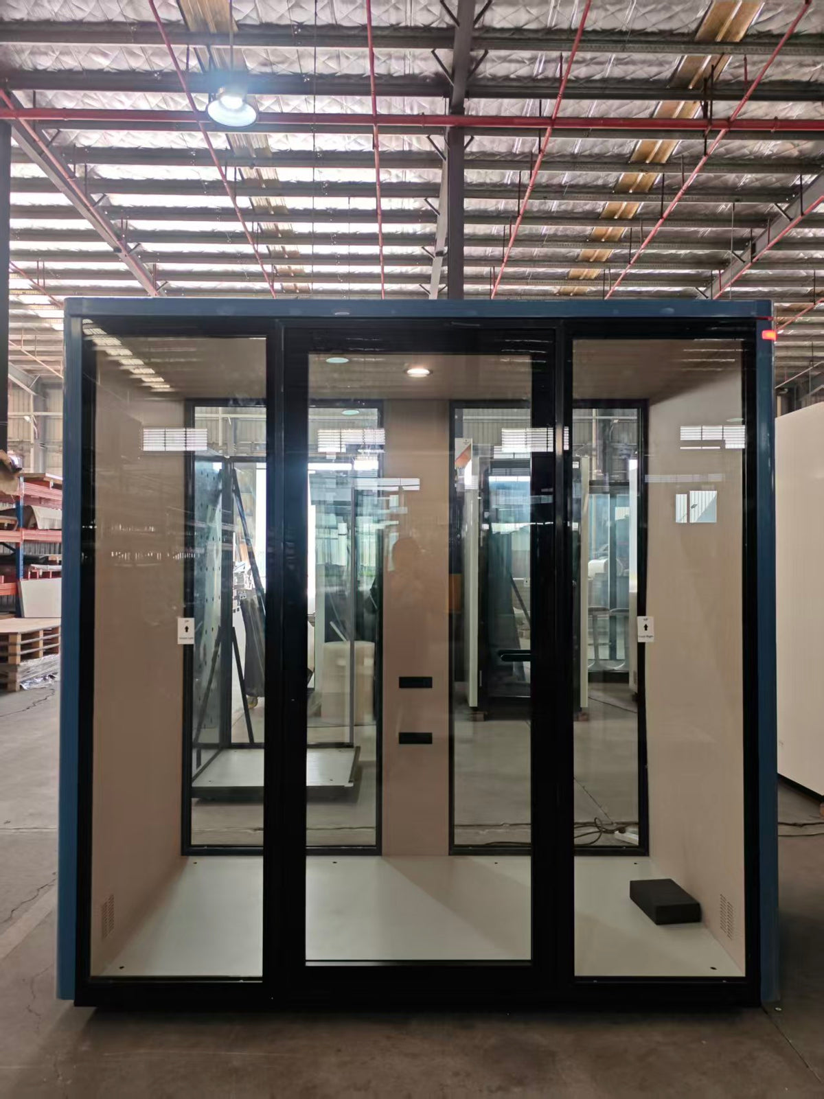 A portable glass partition booth or privacy pod with a black metal frame and beige interior walls, shown in a warehouse showroom.