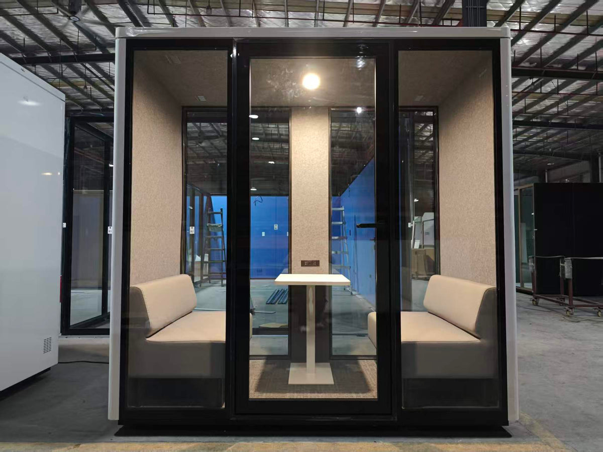 A modern enclosed meeting pod with black-framed glass walls, beige upholstered bench seating on both sides, and a small white table in the center, photographed in a warehouse setting.