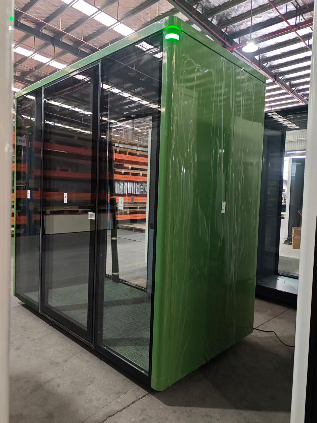 A green enclosed office pod or cabin with large black-framed glass doors, photographed in a warehouse setting. The exterior paneling is wrapped in protective plastic, and a small green indicator light glows near the top edge.