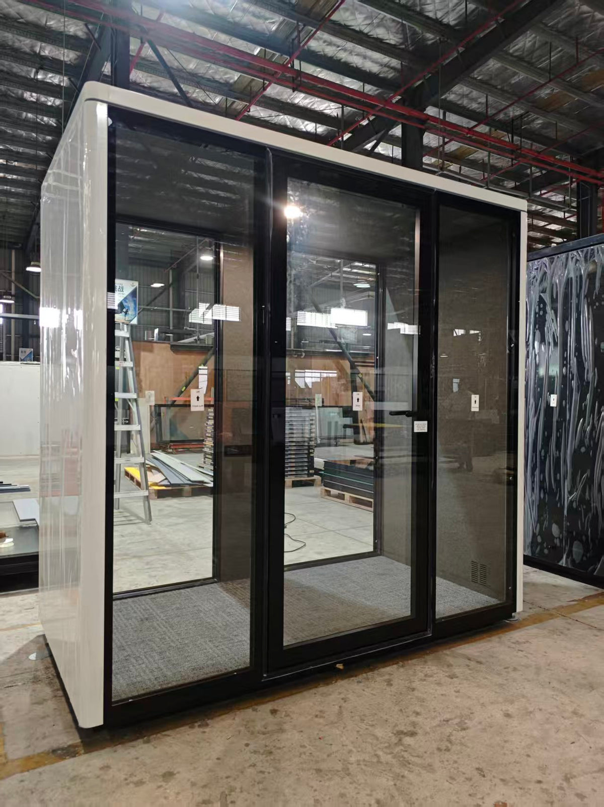 A modern modular office pod with a white exterior and black-framed glass sliding doors, shown in a warehouse setting.