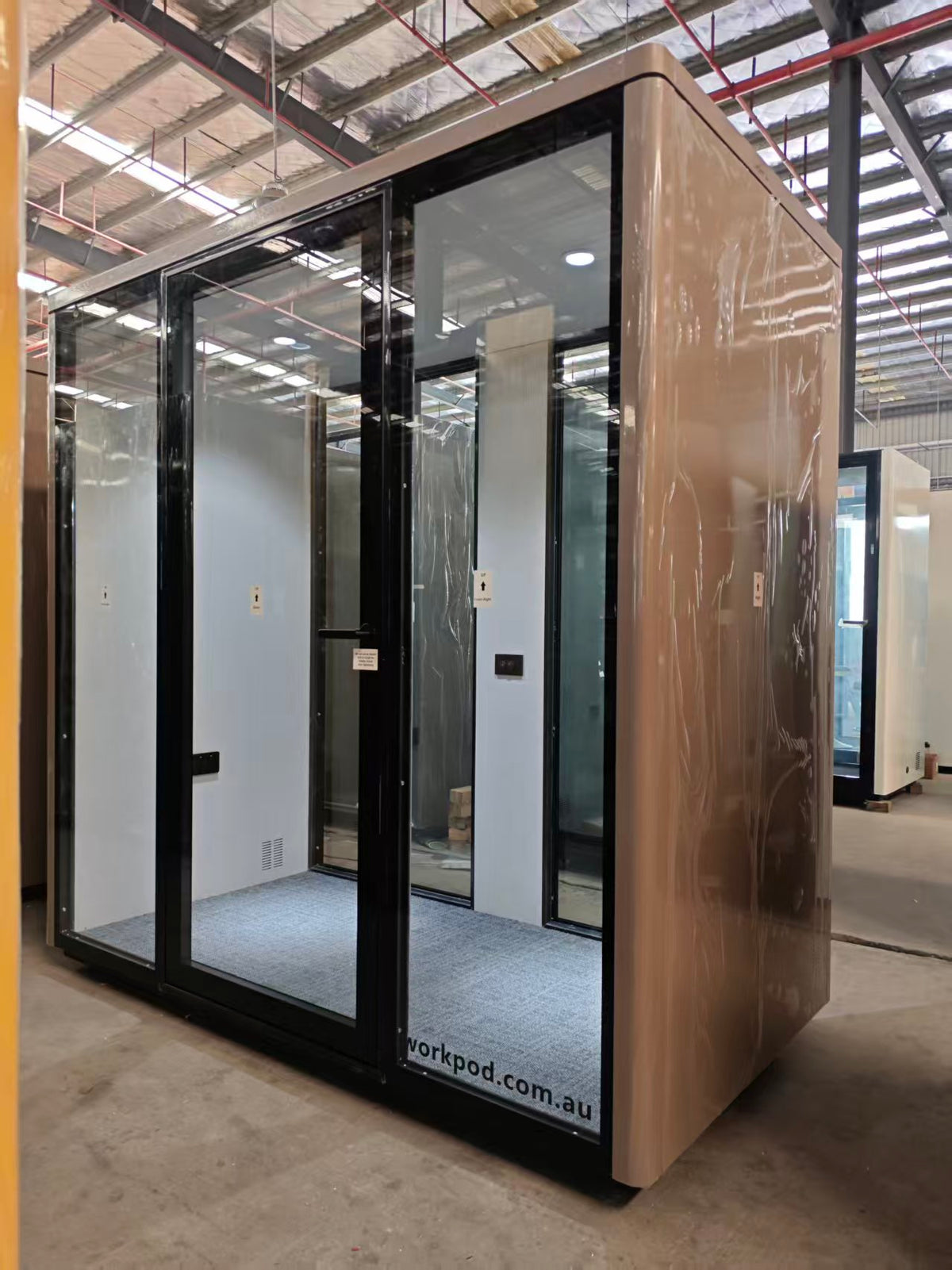 A modern glass office pod or privacy room with a beige side panel and black frame, shown inside a warehouse. The pod has a protective plastic wrap on the exterior and a visible 'workpod.