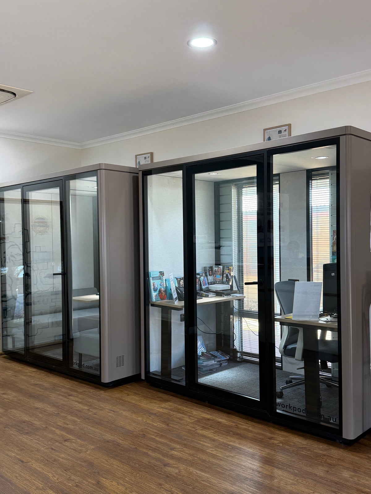 Modern office partition pods with black-framed glass sliding doors and gray side panels, installed in an office space with wood flooring and desks visible inside.