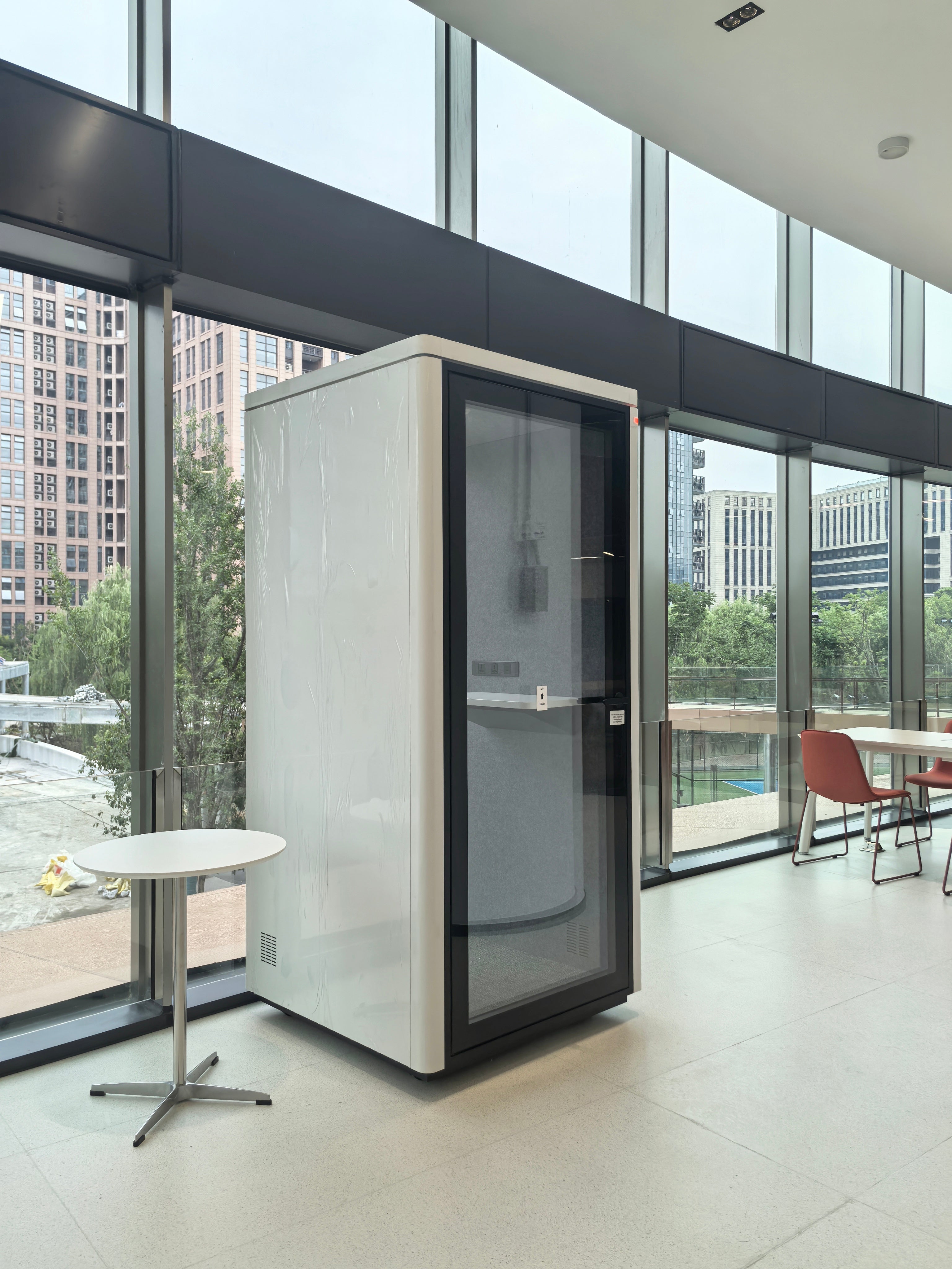 A modern white privacy pod phone booth with a black-framed glass door, set inside a contemporary office space with large windows and city views.