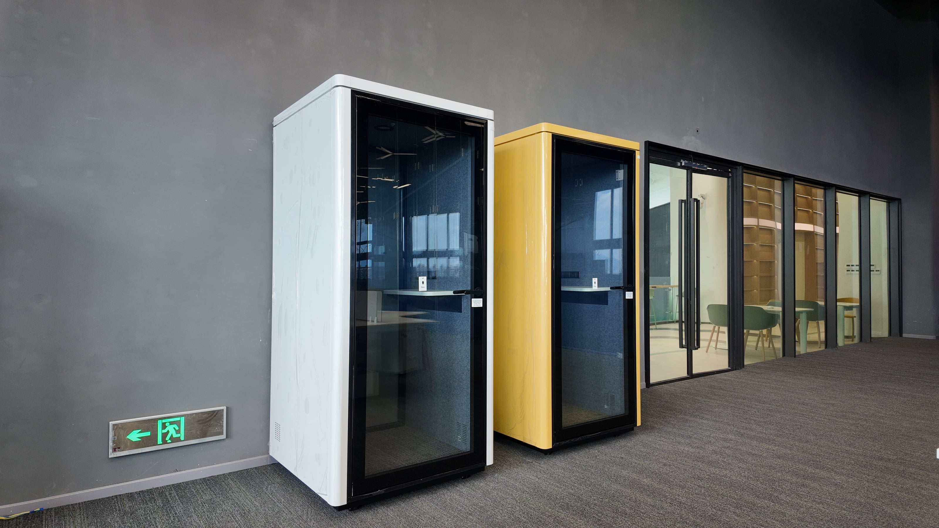 Two modern glass-paneled meeting pods, one white and one yellow, standing in a gray office lobby next to glass conference walls.
