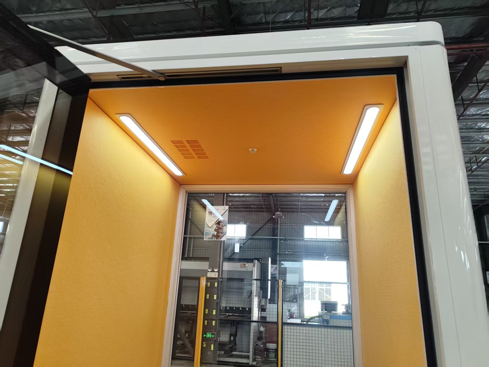 A white industrial machine or enclosure with a bright yellow interior, shown in a warehouse or factory setting. The opening has a black frame, and the interior is illuminated by two long fluorescent lights.