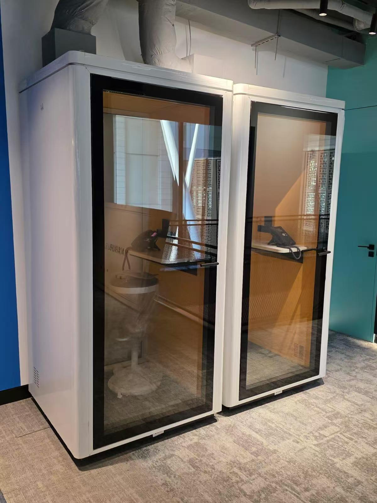 Two white privacy call rooms with large black-framed glass doors, shown in an office-like interior with patterned carpet and a teal wall.