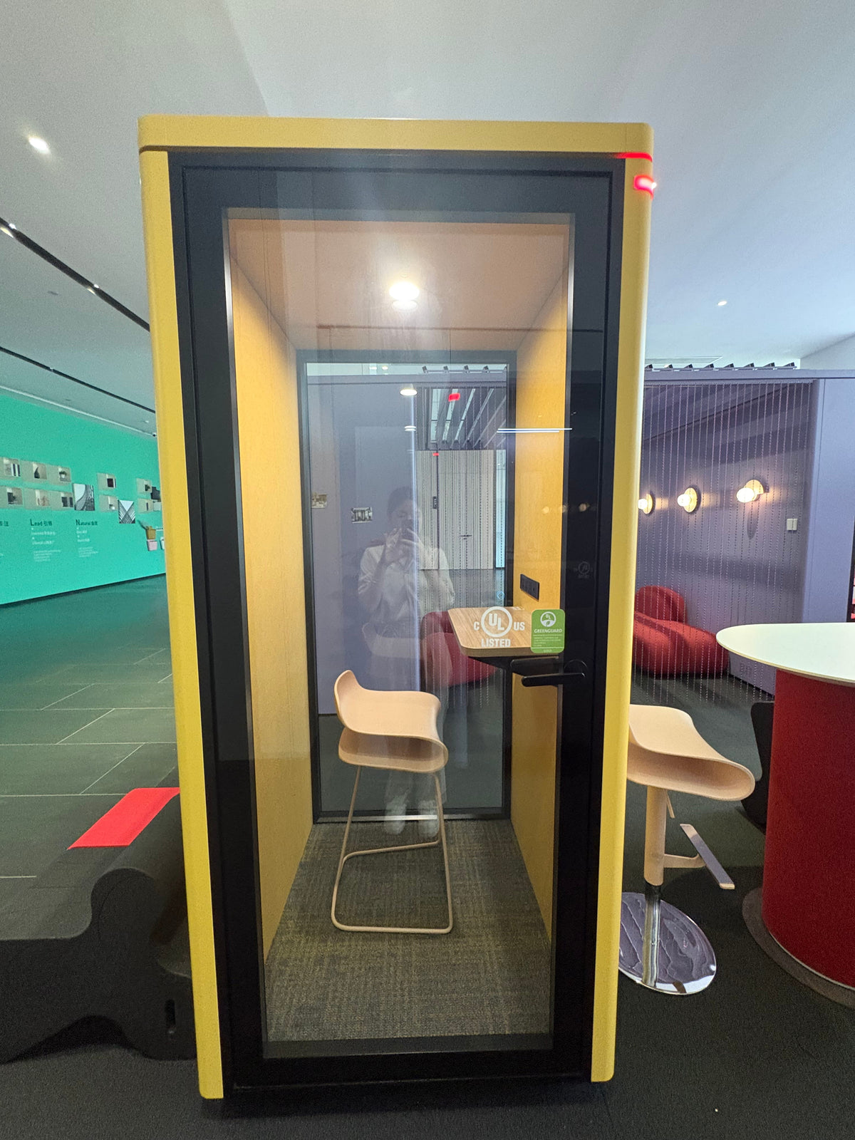 A yellow and black glass conference phone booth with a small table and two beige chairs inside, photographed in a modern office lobby.