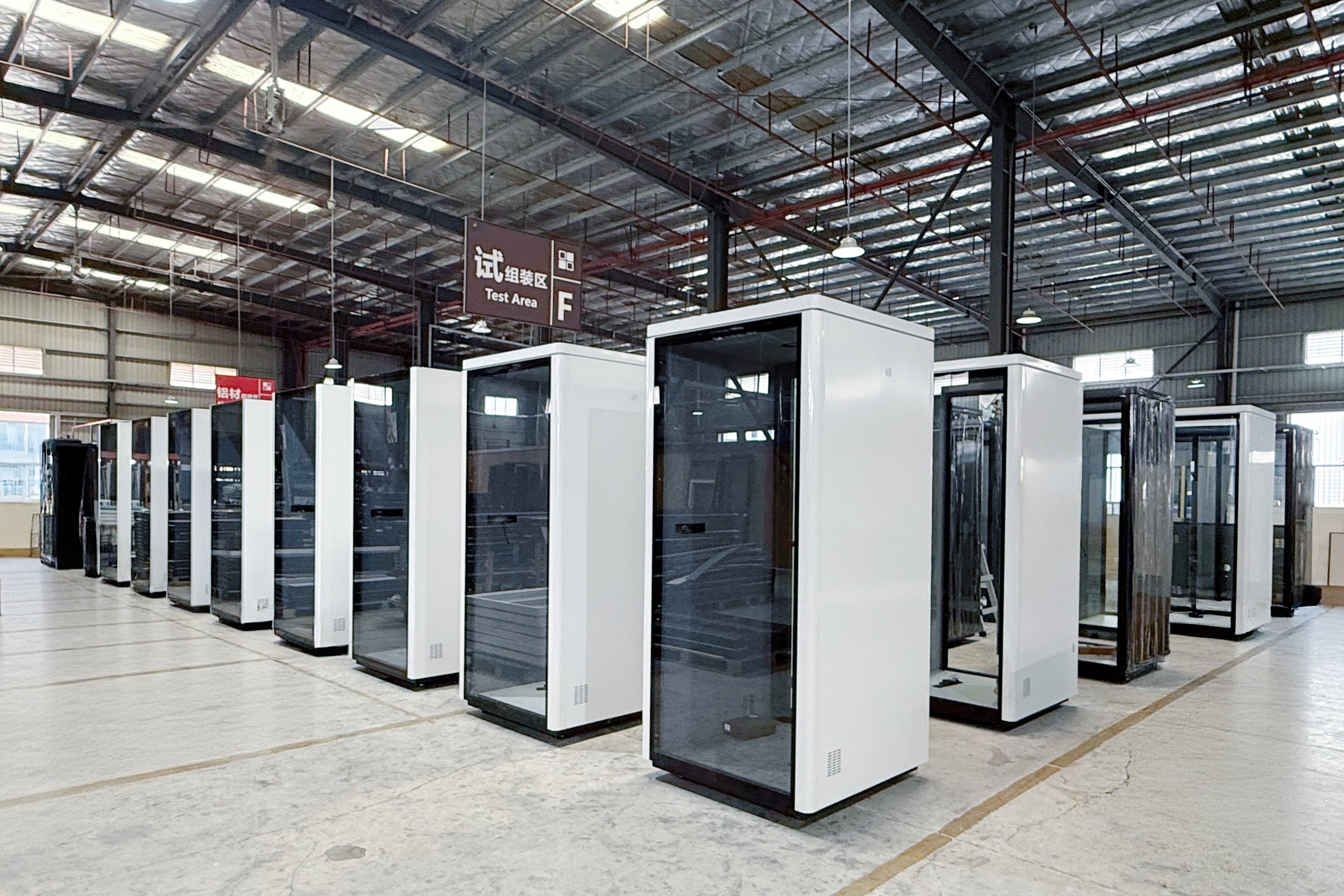 A row of modern white and black glass privacy pods or server closets arranged in a large industrial warehouse with high metal roofs and concrete floors.