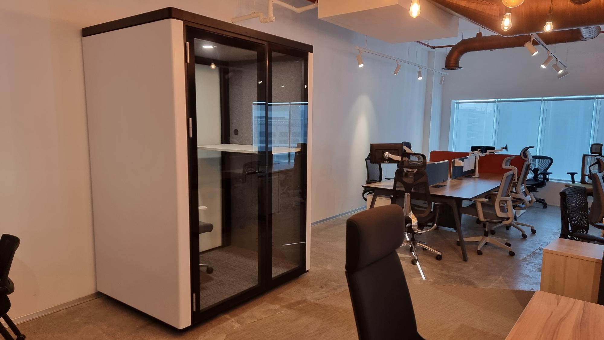 A modern office interior with a large white and black glass partition pod or meeting room on the left, alongside an open-plan workspace with desks, chairs, and windows in the background.