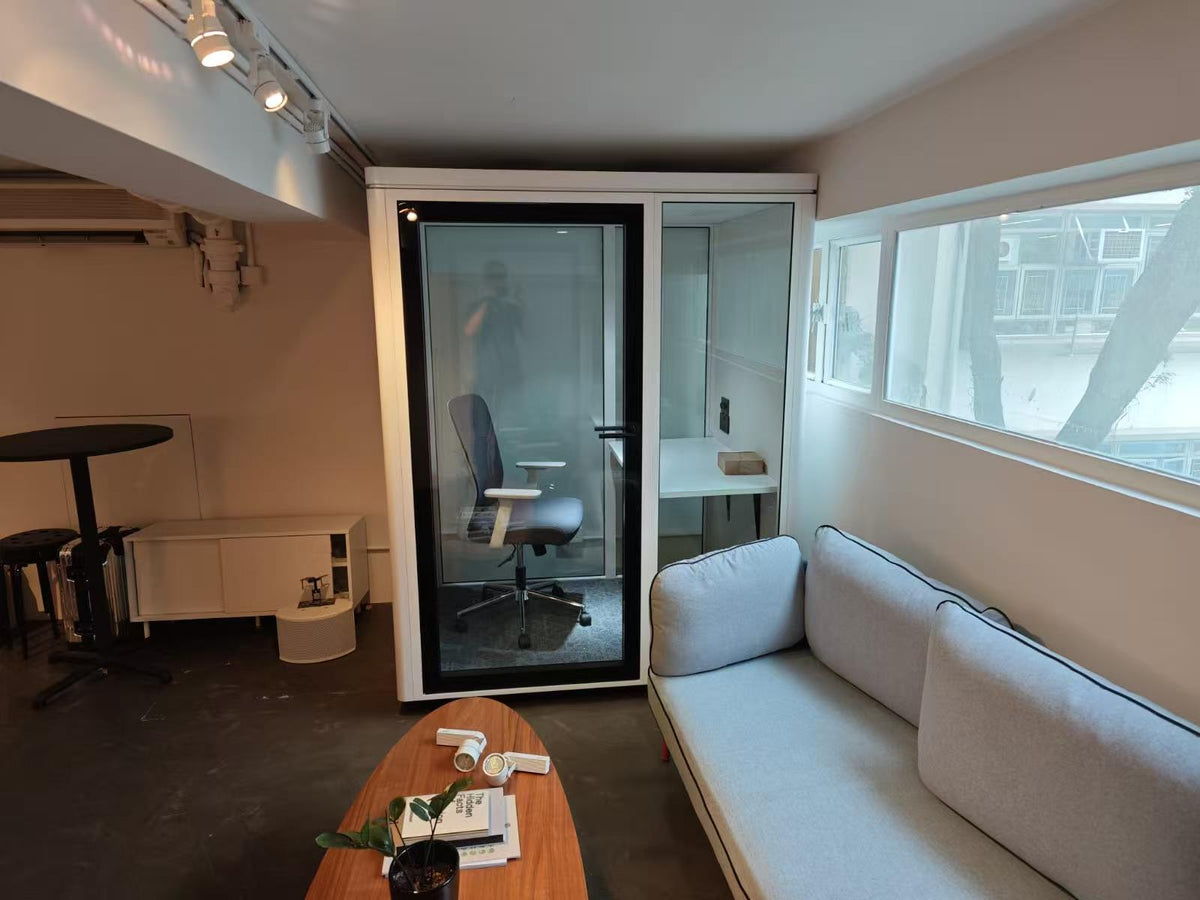 A modern office interior with a light gray fabric sofa in the foreground, a wooden coffee table, and a glass-walled private office booth. The room has beige walls, a dark concrete floor, and a large window on the right.