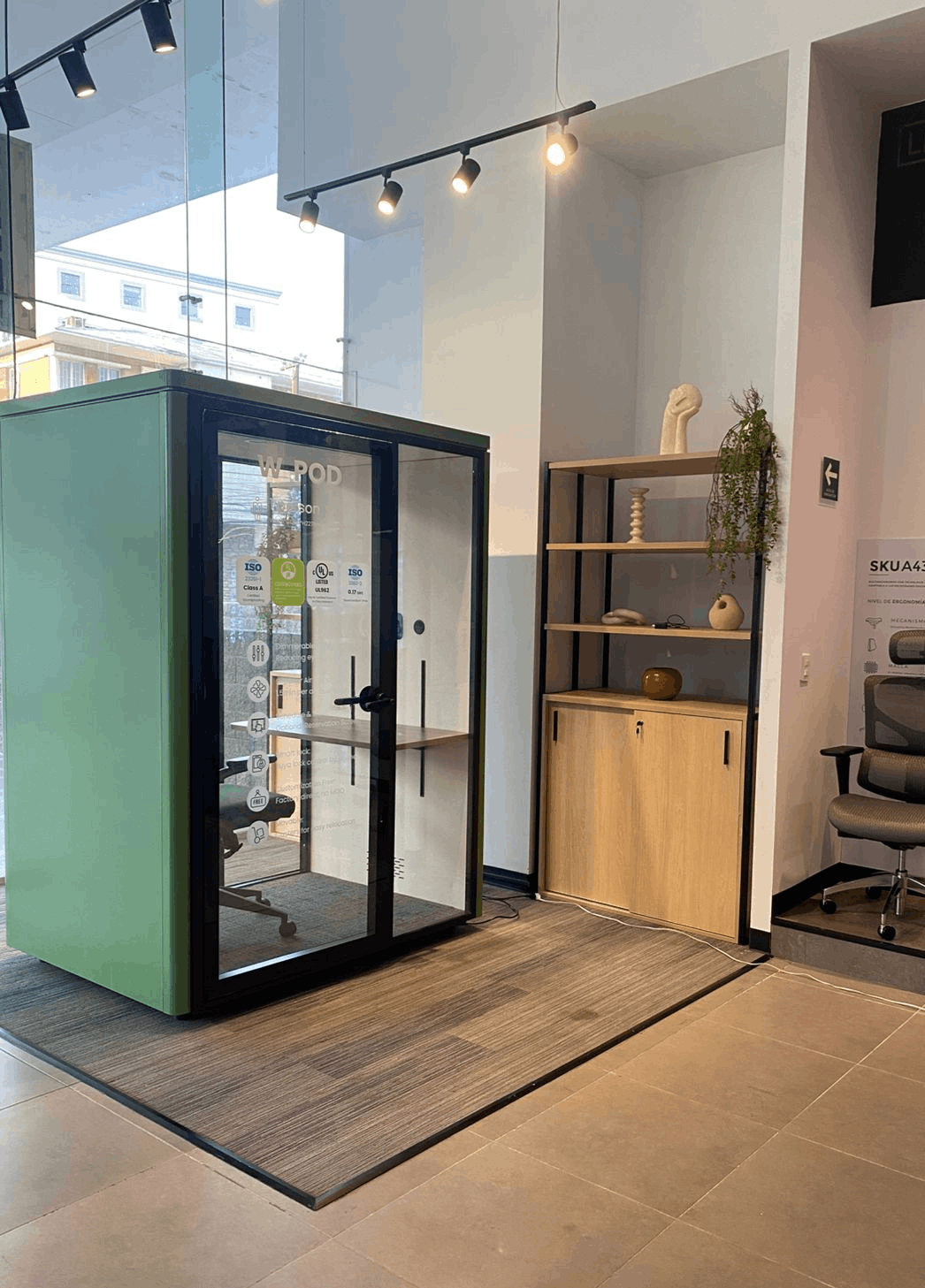 A modern green and black soundproof office pod with glass doors, shown in a showroom next to a wooden shelving unit and office chair.