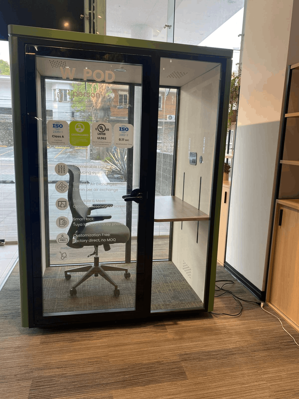 A W-POD glass office pod with a black frame, shown in a showroom. The pod features a glass door with a black handle, a built-in desk shelf, and a gray office chair inside.