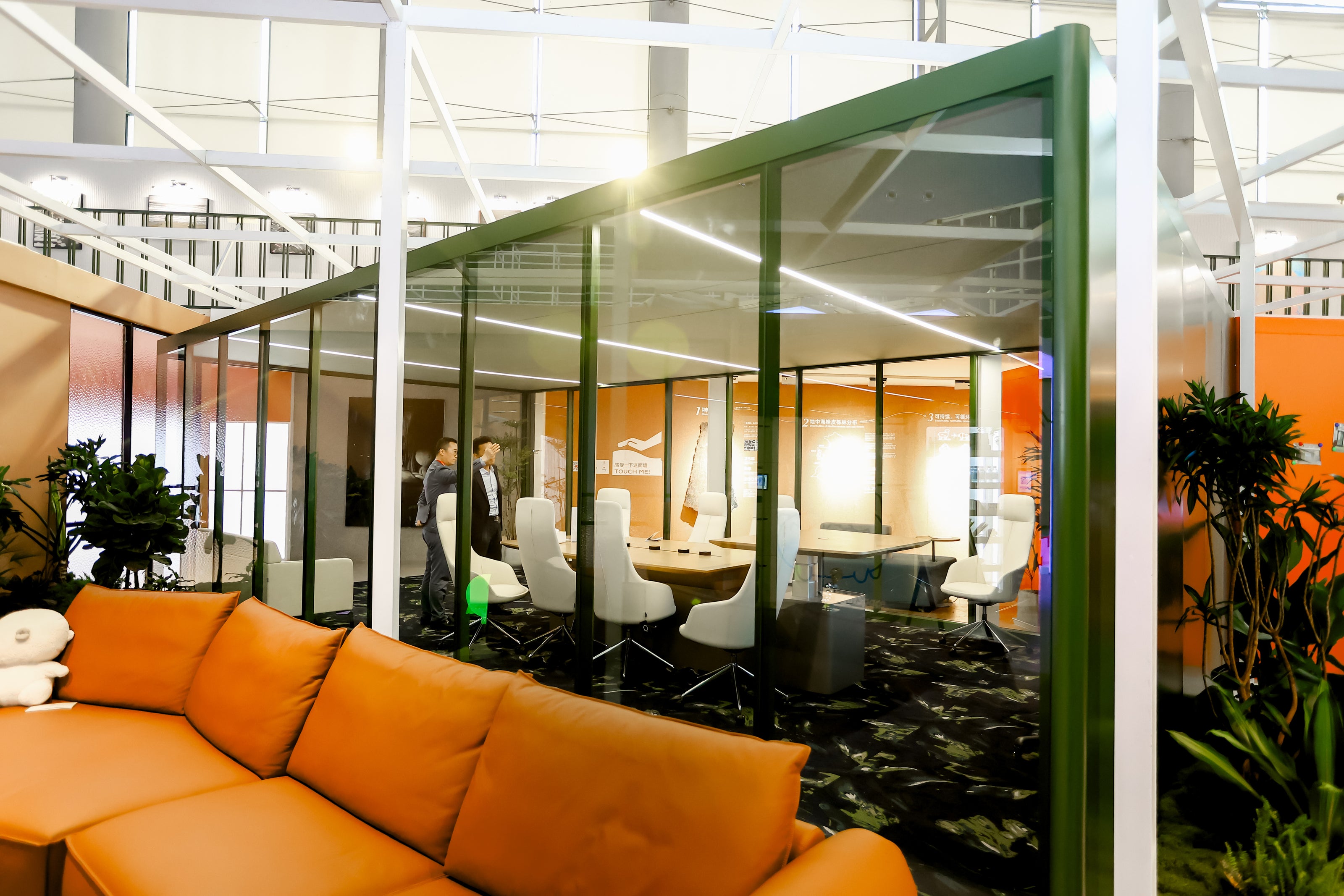 Modern room-in-room office space with glass partition, orange sofa, and people in the background.