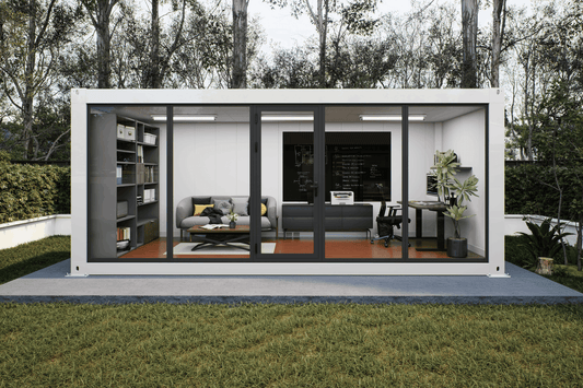 A modern white modular office or container home with large black-framed glass doors, shown open with furnished interior rooms including a gray sofa, desk, shelving, and office setup. The structure sits on a concrete pad in a grassy yard with trees behind it.