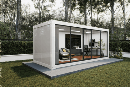 A modern white shipping container home with large glass sliding doors, shown on a concrete platform in a grassy backyard with trees in the background. The interior is furnished with a gray sofa, office desk, and chair.