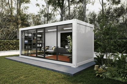 A white modular office container home with large glass windows, shown on a concrete platform in a landscaped garden with trees and grass.