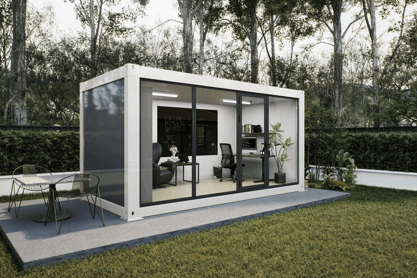 A modern white modular office pod with large black-framed glass windows, set on a concrete patio in a grassy backyard with trees in the background. The office interior is furnished with a desk, office chair, and shelving, and an outdoor dining table is visible on the patio.