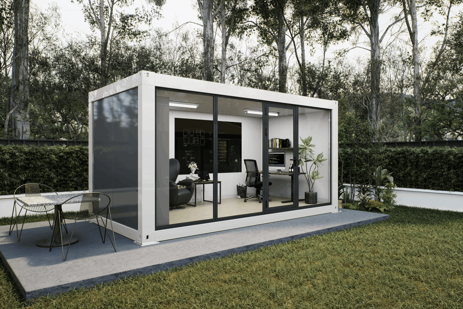 A modern white modular office pod with large black-framed glass windows, set on a concrete patio in a grassy backyard with trees in the background. The office interior is furnished with a desk, office chair, and shelving, and an outdoor dining table is visible on the patio.
