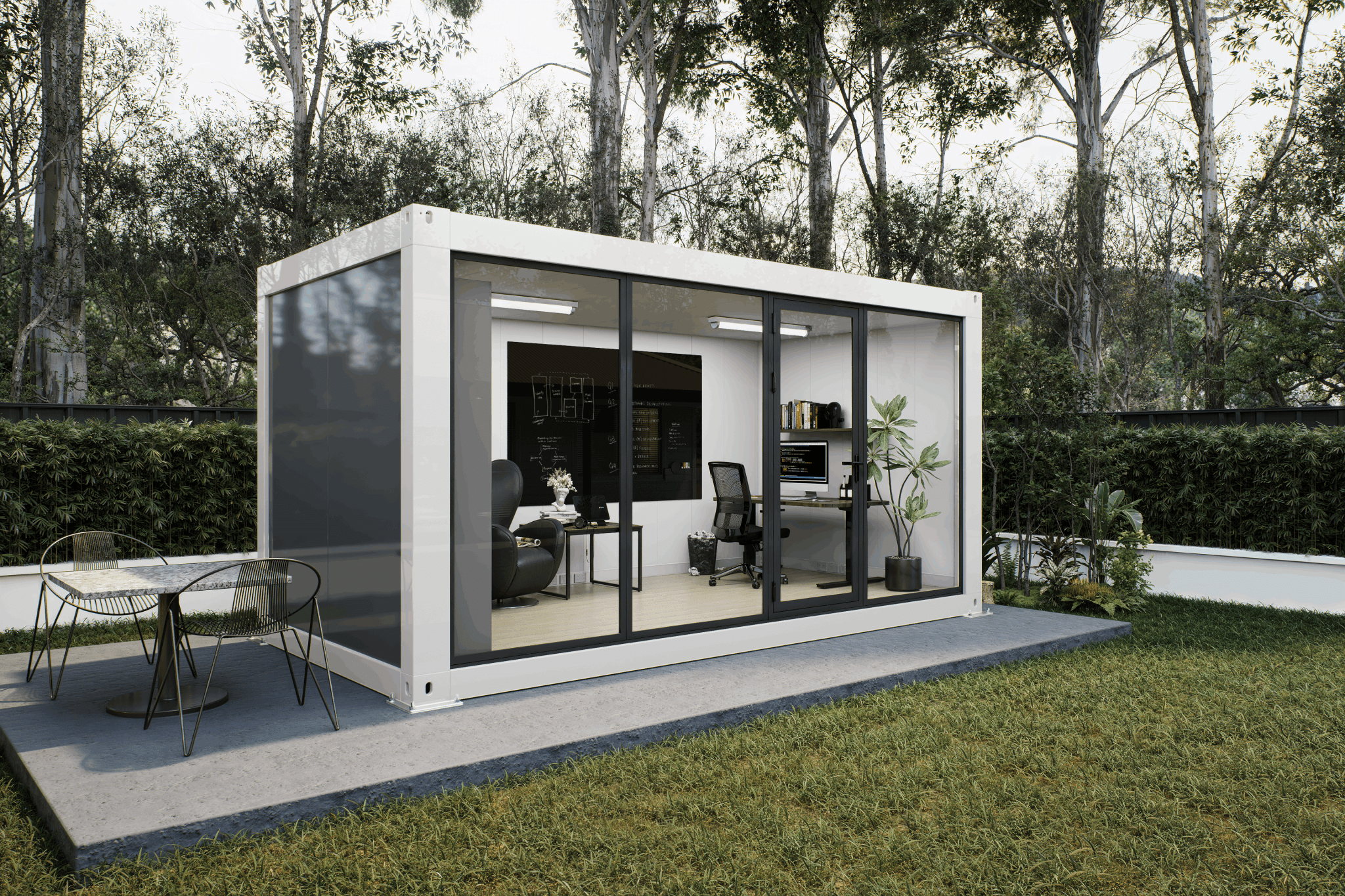 A modern white modular office pod with large black-framed glass windows, set on a concrete patio in a grassy backyard with trees in the background. The office interior is furnished with a desk, office chair, and shelving, and an outdoor dining table is visible on the patio.