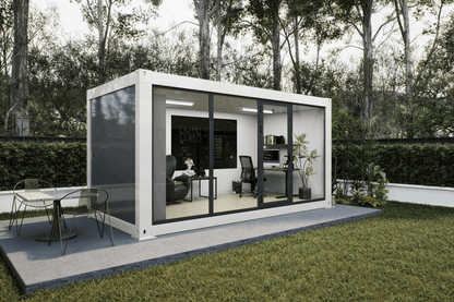 A modern white modular office pod with large black-framed glass windows, set on a concrete patio in a grassy backyard with trees in the background. The office interior is furnished with a desk, office chair, and shelving, and an outdoor dining table is visible on the patio.