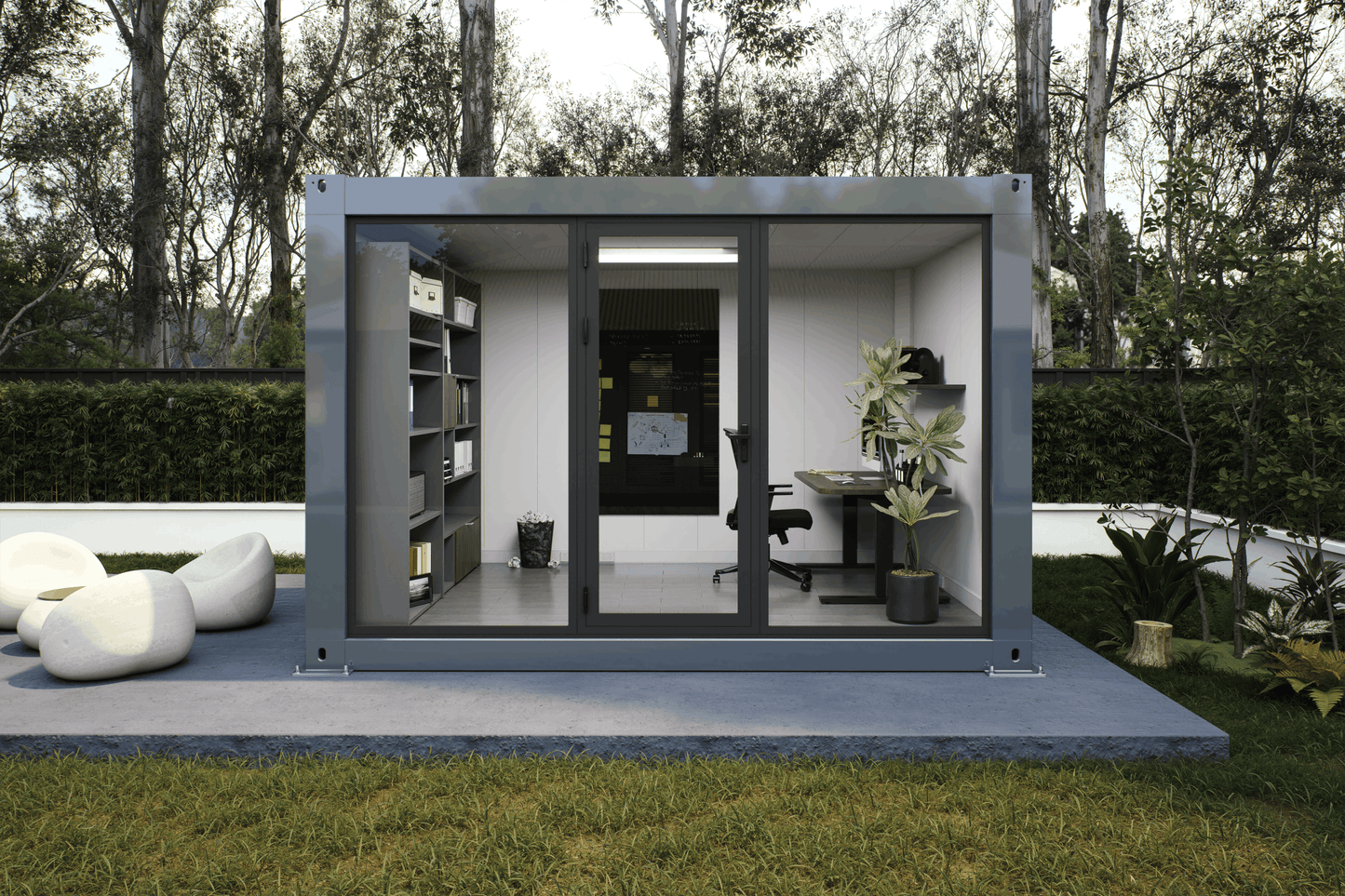 A modern modular office pod with a gray metal frame and glass walls, set on a concrete patio in a grassy backyard with trees in the background. The office interior features a desk, office chair, shelving, and plants.
