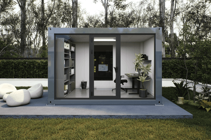 A modern modular office pod with a gray metal frame and glass walls, set on a concrete patio in a grassy backyard with trees in the background. The office interior features a desk, office chair, shelving, and plants.
