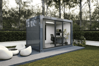A modern modular office pod with a dark gray metal frame and glass sliding doors, situated on a concrete patio in a landscaped garden with green grass, hedges, and trees.