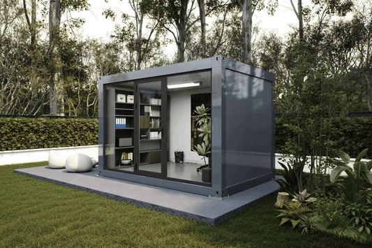A modern gray modular outdoor office pod with large glass windows, shown on a concrete platform in a landscaped garden with trees and grass.