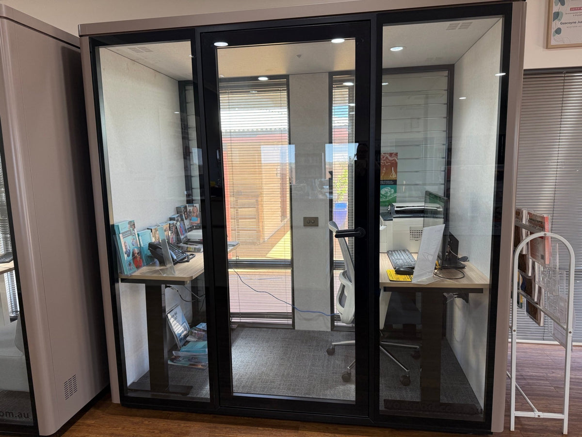 A glass office partition wall with black metal frames, enclosing a small workspace with a desk, chair, and computer setup. The room is shown in a real office environment with additional furniture and blinds visible.