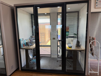 A glass office partition wall with black metal frames, enclosing a small workspace with a desk, chair, and computer setup. The room is shown in a real office environment with additional furniture and blinds visible.