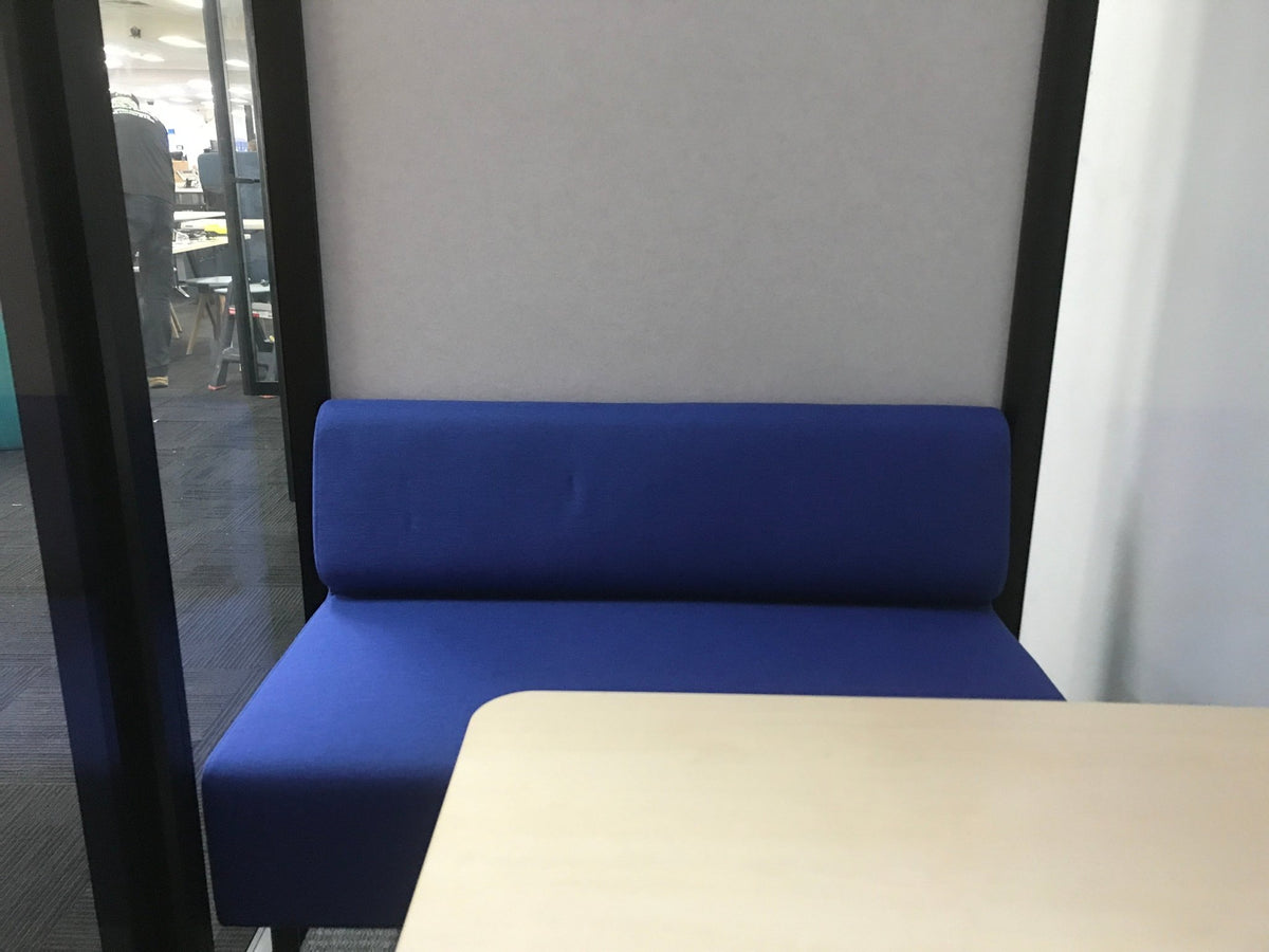 A blue upholstered booth or bench seat with a tall backrest, shown in an office-like interior with a light wood table in the foreground and a gray wall behind it.