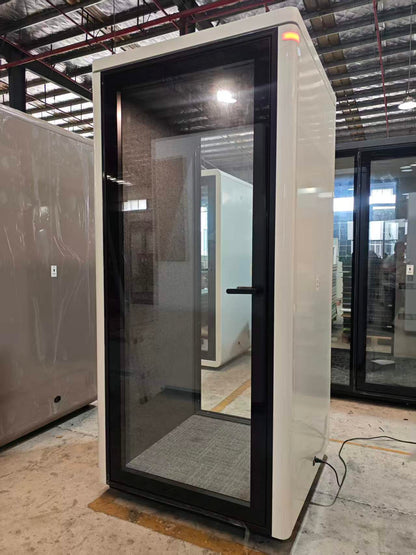 A modern outdoor privacy booth with a white exterior and black-framed glass doors, shown in a warehouse setting.