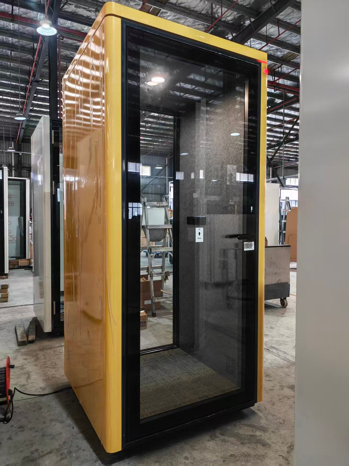 A modern enclosed privacy booth with a glossy mustard yellow exterior and a black-framed glass door, photographed in a warehouse setting.