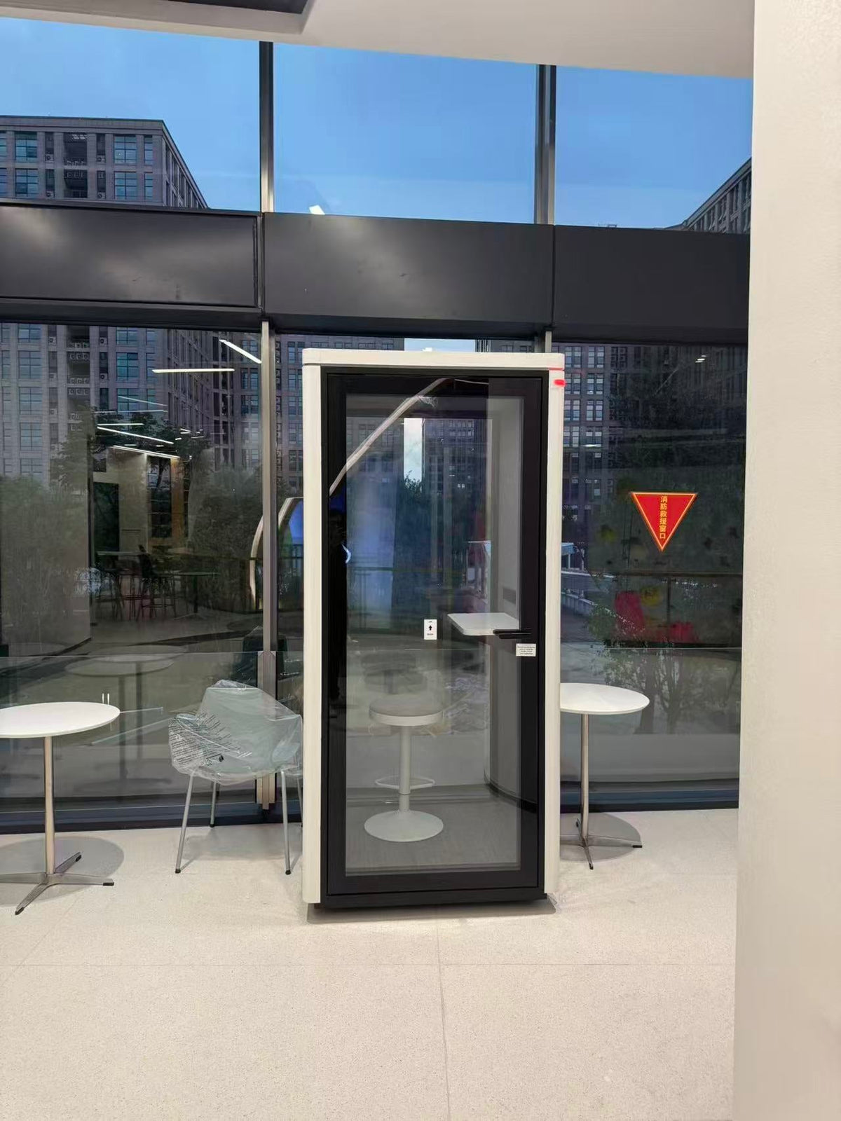 A modern glass and black metal privacy booth or call center enclosure standing indoors in front of large floor-to-ceiling windows. The unit has a white outer frame, a black central frame, a glass door panel, a small white step platform at the top, and a vertical handle on the left side.