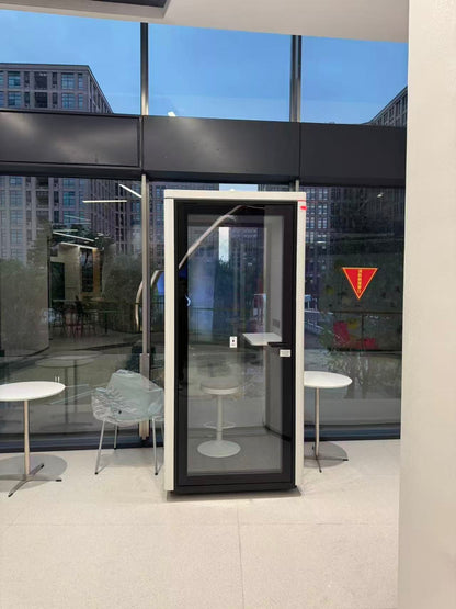 A modern glass and black metal privacy booth or call center enclosure standing indoors in front of large floor-to-ceiling windows. The unit has a white outer frame, a black central frame, a glass door panel, a small white step platform at the top, and a vertical handle on the left side.