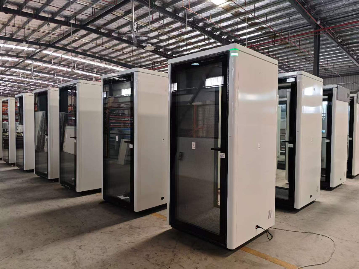 A row of white and black privacy booths with glass doors, displayed inside a large warehouse or factory with a high metal roof.