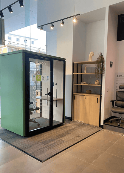 A green and black enclosed office phone booth with glass doors is shown in a showroom interior next to a wooden shelving unit and office chair.