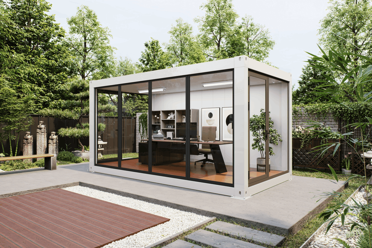 A modern white shipping container office pod with large black-framed glass windows, situated on a concrete patio in a lush green backyard setting.