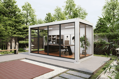 A modern white shipping container office pod with large black-framed glass windows, situated on a concrete patio in a lush green backyard setting.