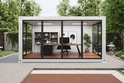 A modern white modular office pod with large black-framed glass doors, set in a landscaped backyard with trees, a concrete patio, and a wooden deck. Inside, a home office setup with a desk, chair, shelves, and computer is visible.