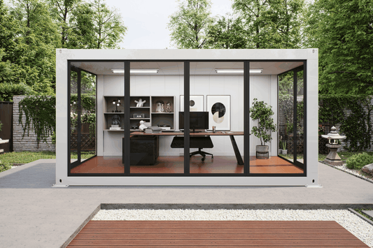 A modern white modular office pod with large black-framed glass doors, set in a landscaped backyard with trees, a concrete patio, and a wooden deck. Inside, a home office setup with a desk, chair, shelves, and computer is visible.