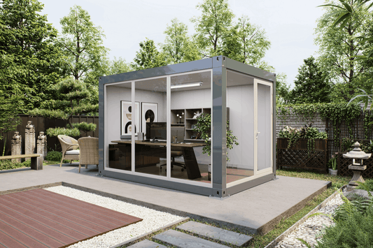 A modern modular office pod with a gray metal frame and glass walls, situated on a concrete patio in a landscaped garden setting.