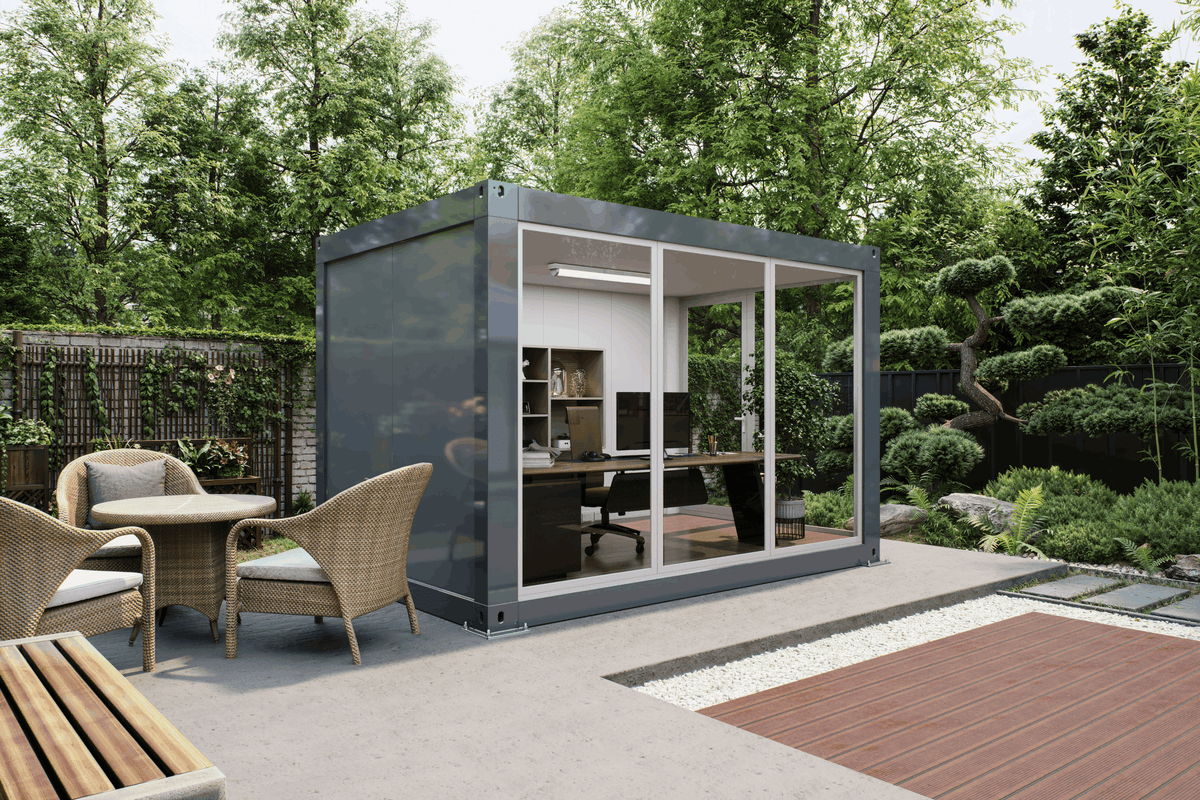 A modern gray shipping container office pod with large glass walls, situated on a concrete patio in a landscaped garden setting with outdoor dining chairs and a wooden deck.