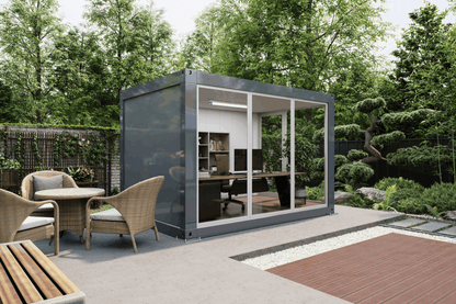 A modern gray shipping container office pod with large glass walls, situated on a concrete patio in a landscaped garden setting with outdoor dining chairs and a wooden deck.