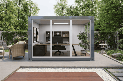 A modern modular office pod with a gray metal frame and glass walls, situated on a paved patio in a landscaped garden setting. Inside the pod, a home office setup with a desk, chair, and shelving is visible.